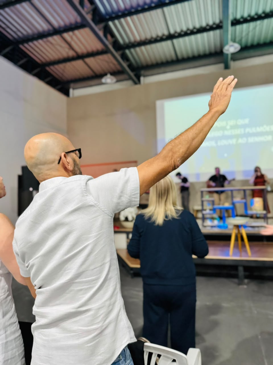 Homem com o braço erguido em adoração durante o louvor, com a equipe ministrando ao fundo.